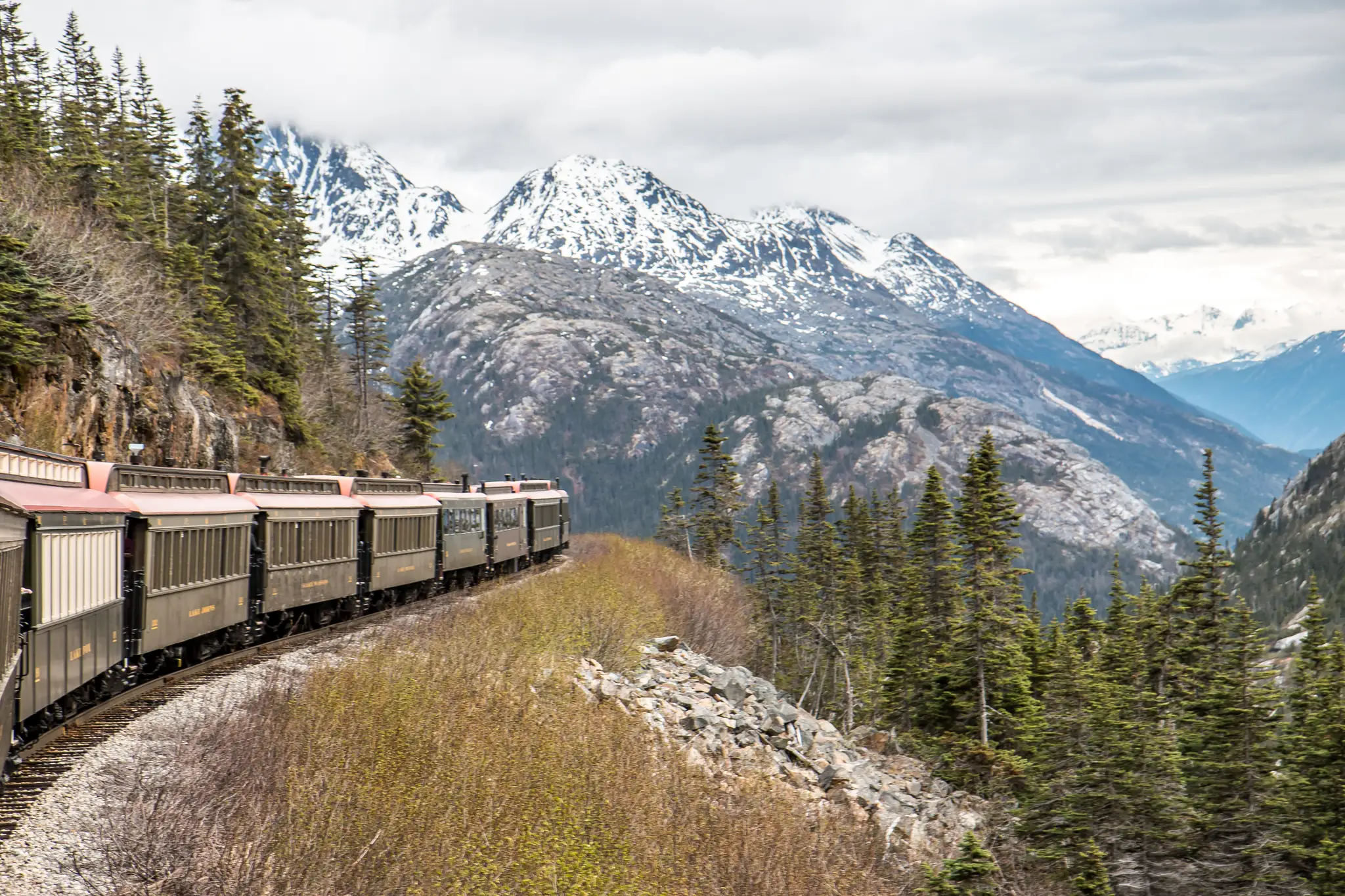 White Pass & Yukon Route train with vintage red-roofed coaches curving along a forested mountainside below snow-capped peaks