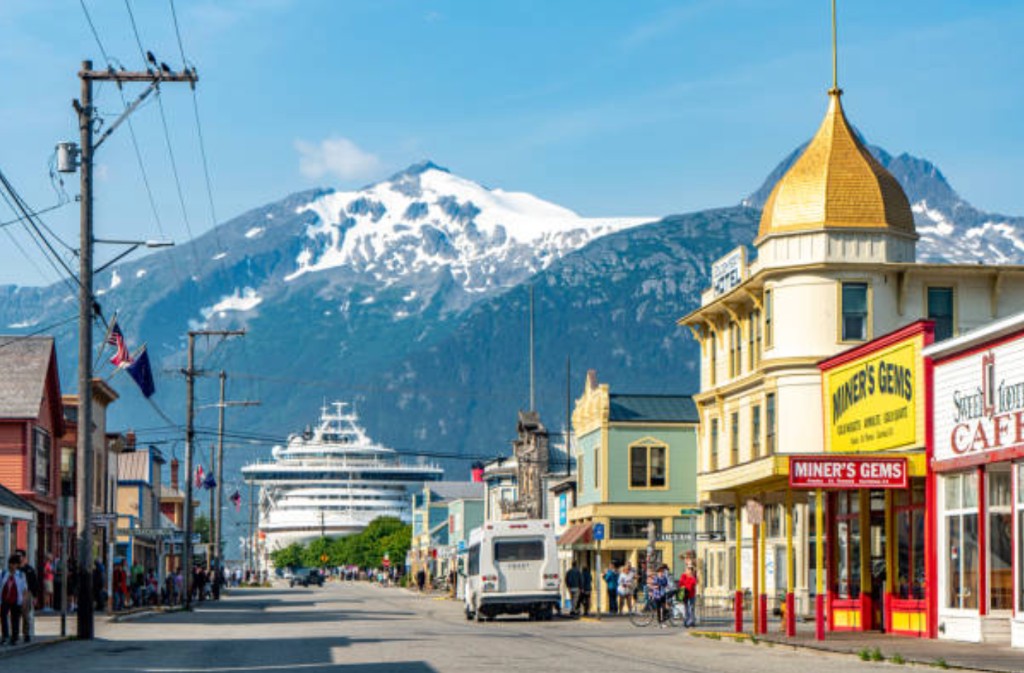 Sunny view of historic Broadway Street in Skagway, Alaska, with colorful Gold Rush buildings, a cruise ship at the pier, and snow-capped mountains