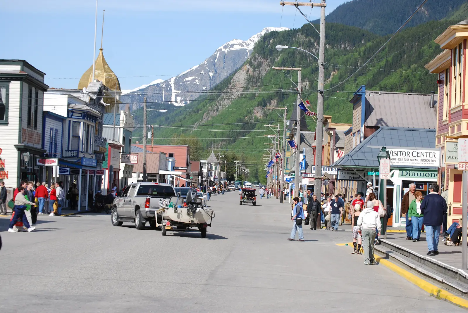 Broadway in Skagway's historic district on a sunny day, with Gold Rush-era storefronts, the gold dome of the Golden North Hotel, and snow-capped mountains beyond