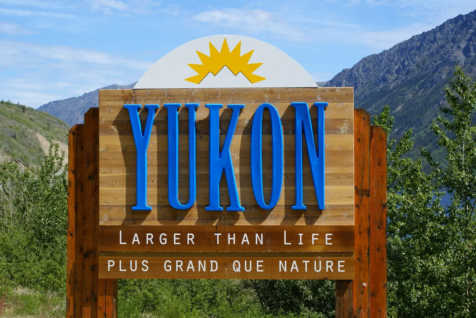 Wooden "Yukon — Larger than Life" welcome sign at the Yukon border, with mountains and blue sky behind